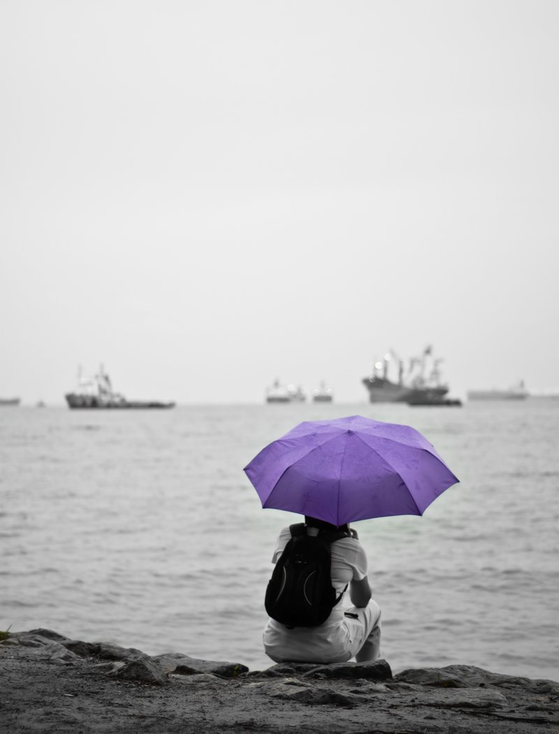 Yksinäinen ihminen istuu kalliolla meren rannalla lila sateenvarjo  kädessään.